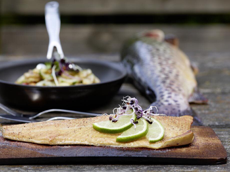 Regionaler und zubereiterter Fisch liegt auf einem Holzbrett, dahinter ist eine Pfanne und ein nicht zubereiteter Fisch.