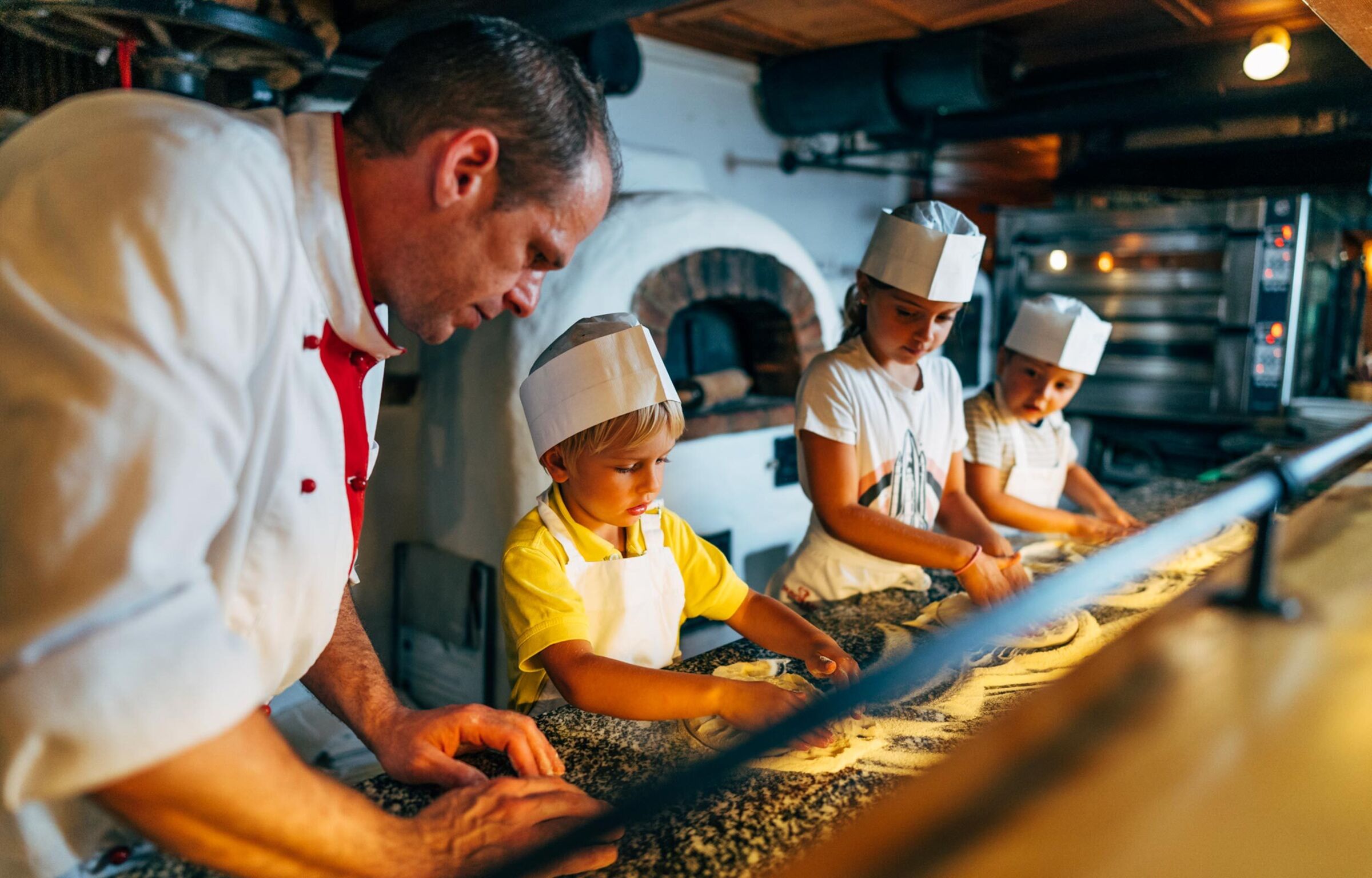 Koch hilft Kinder bei dem kneten des Pizzateiges.