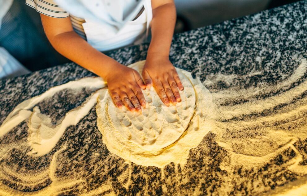 Ein Kind kneten und rollt den Pizzateig mit den Händen und viel Mehl aus