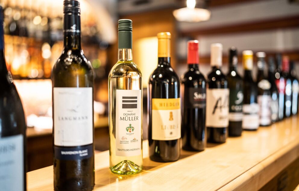 Sul bancone del bar si trovano diverse bottiglie di vino rosso e bianco di diverse cantine e viticoltori.