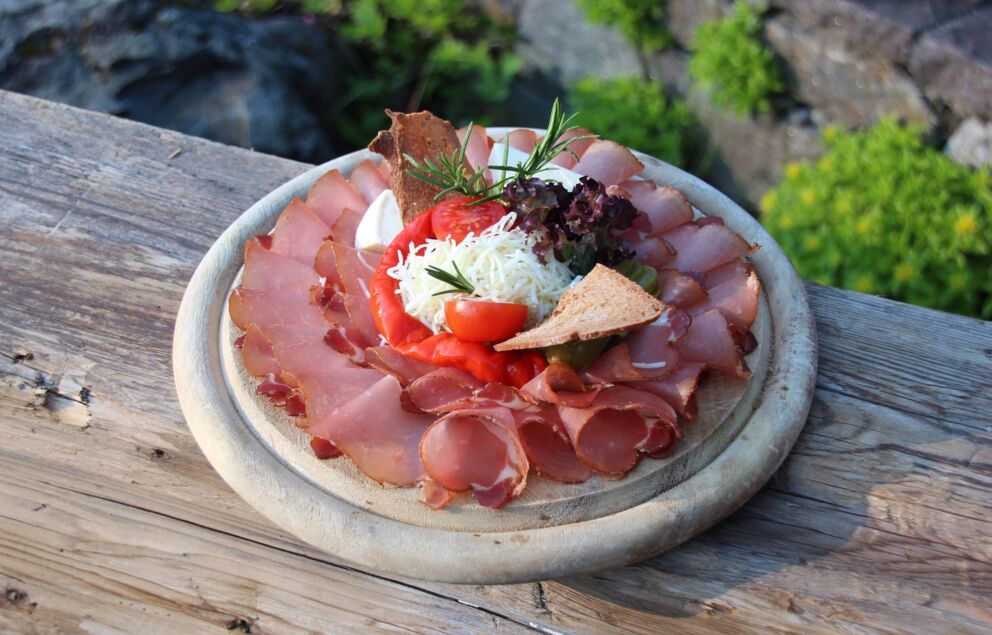 Speck mit Kren, Brot und Pfefferoni ligt auf einem Holzbrett.