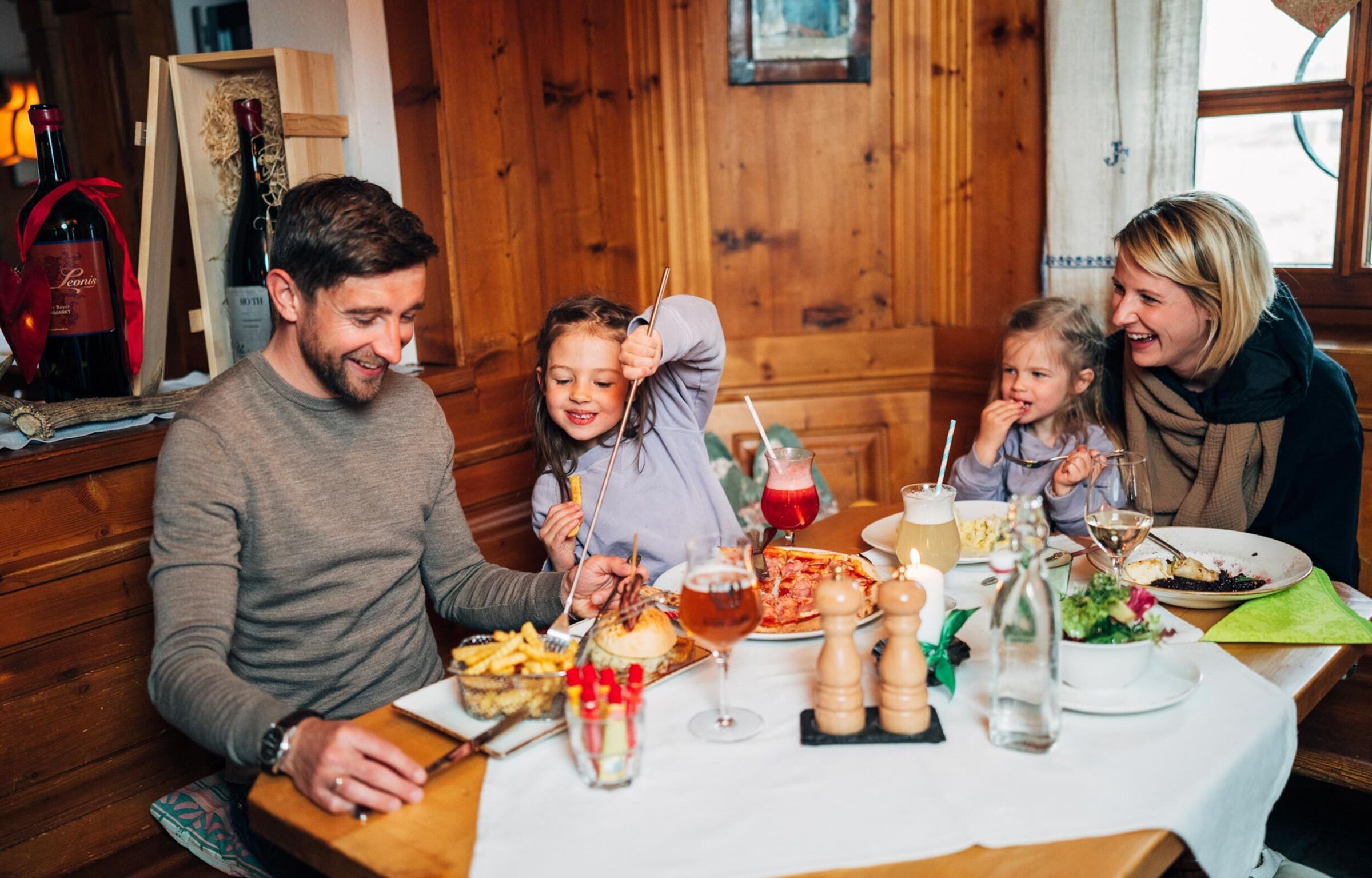 Familie sitzt in einer rustikalen Hütte und essen, eine Tochter klaut dem Vater mit einer Teleskopgabel eine Pommes