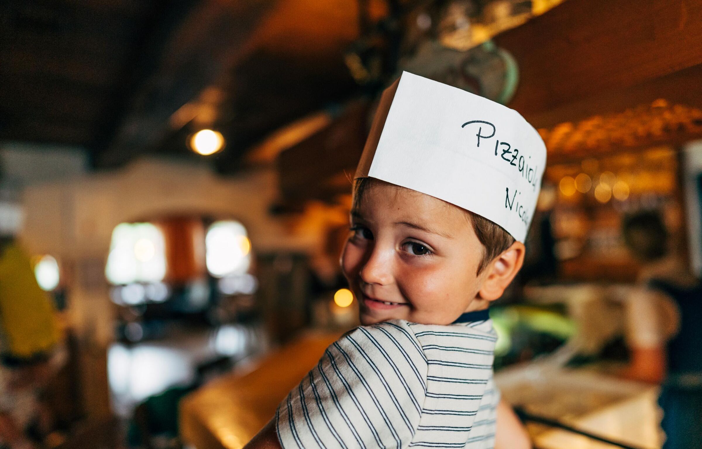 Il bambino ha un cappello da cuoco con la scritta "corso di pizza" e sorride alla telecamera.