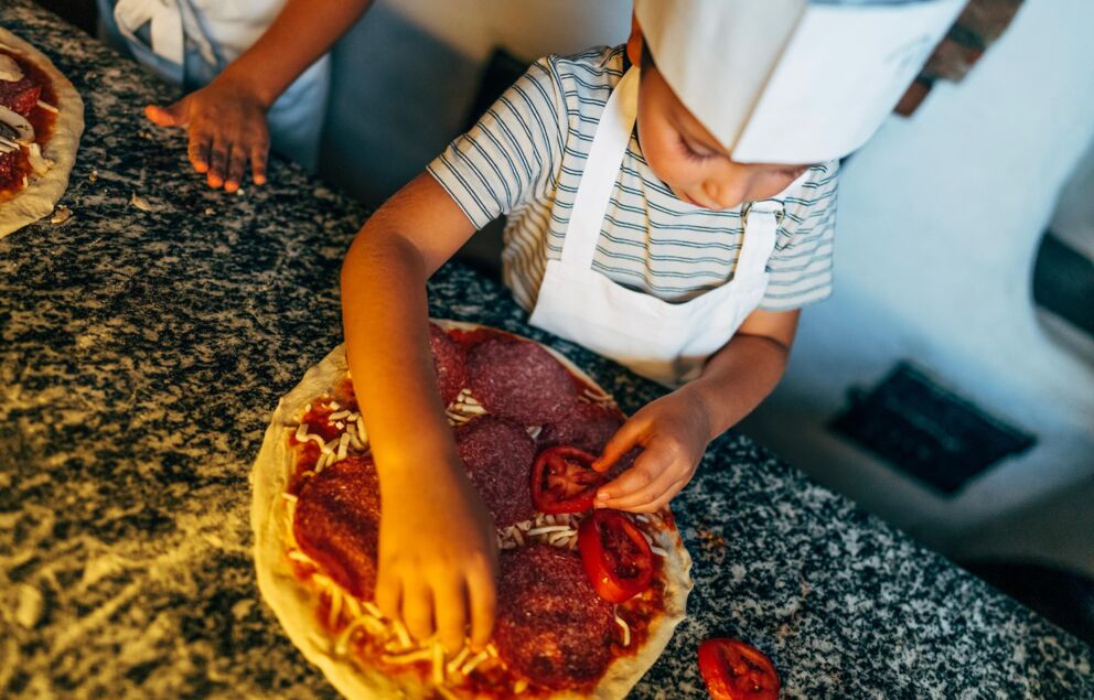 A child in a chef's hat puts different toppings on a pizza