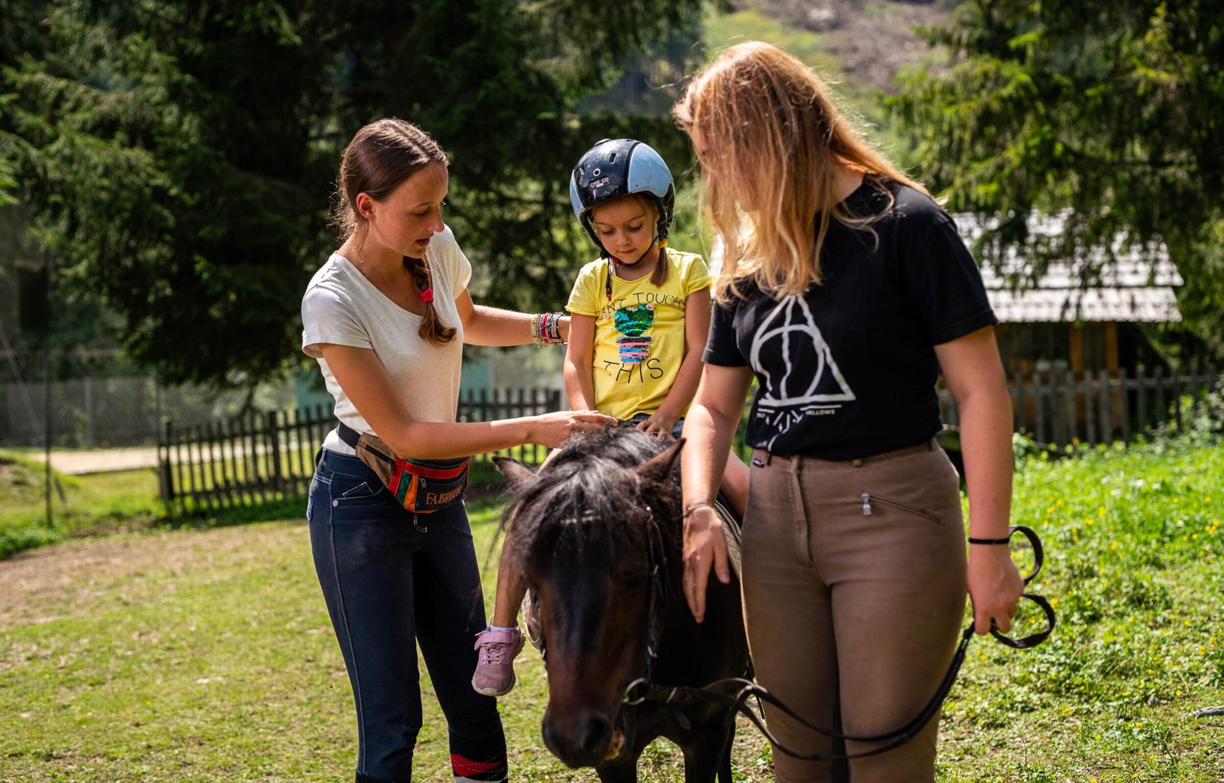 A little girl with a helmet sits on a dark pony and is supported by a woman