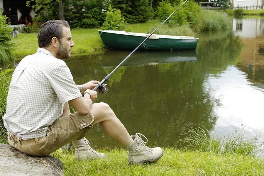 A man in traditional costume sits next to a pond and tries to catch some fish.