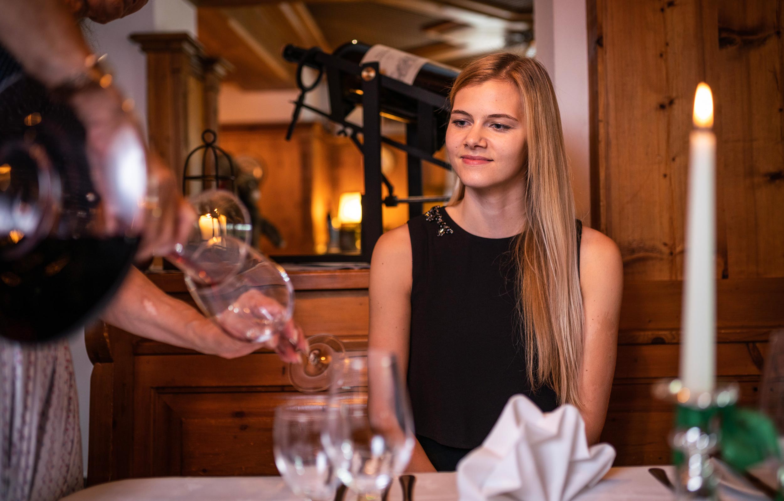 A woman with blond long hair watches a person pouring red wine into a wine glass.