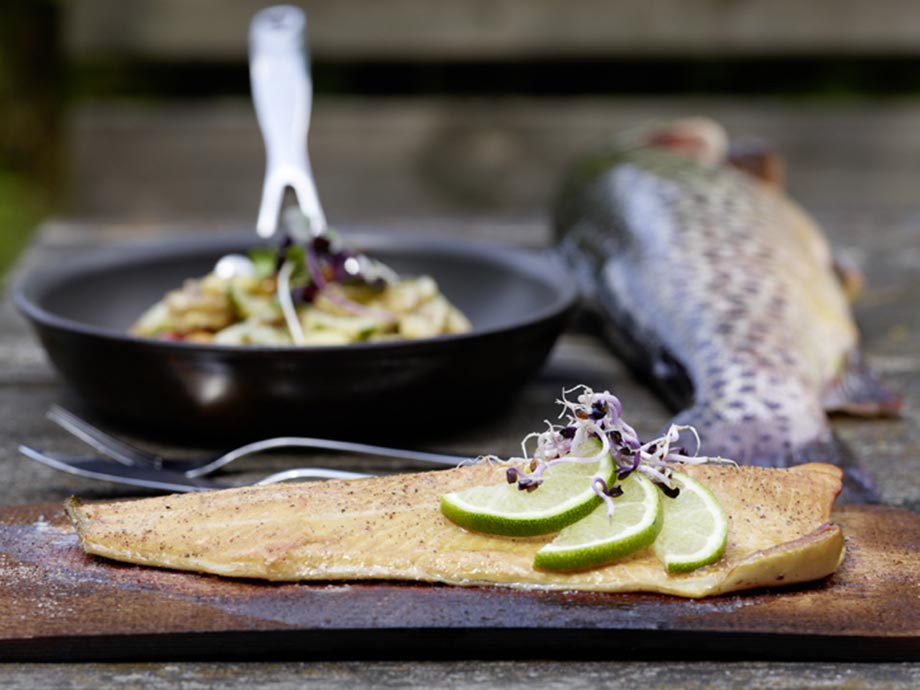 A prepared fish lies on a wooden board garnished with limes, a pan stands behind it and a fresh fish lies next to it.