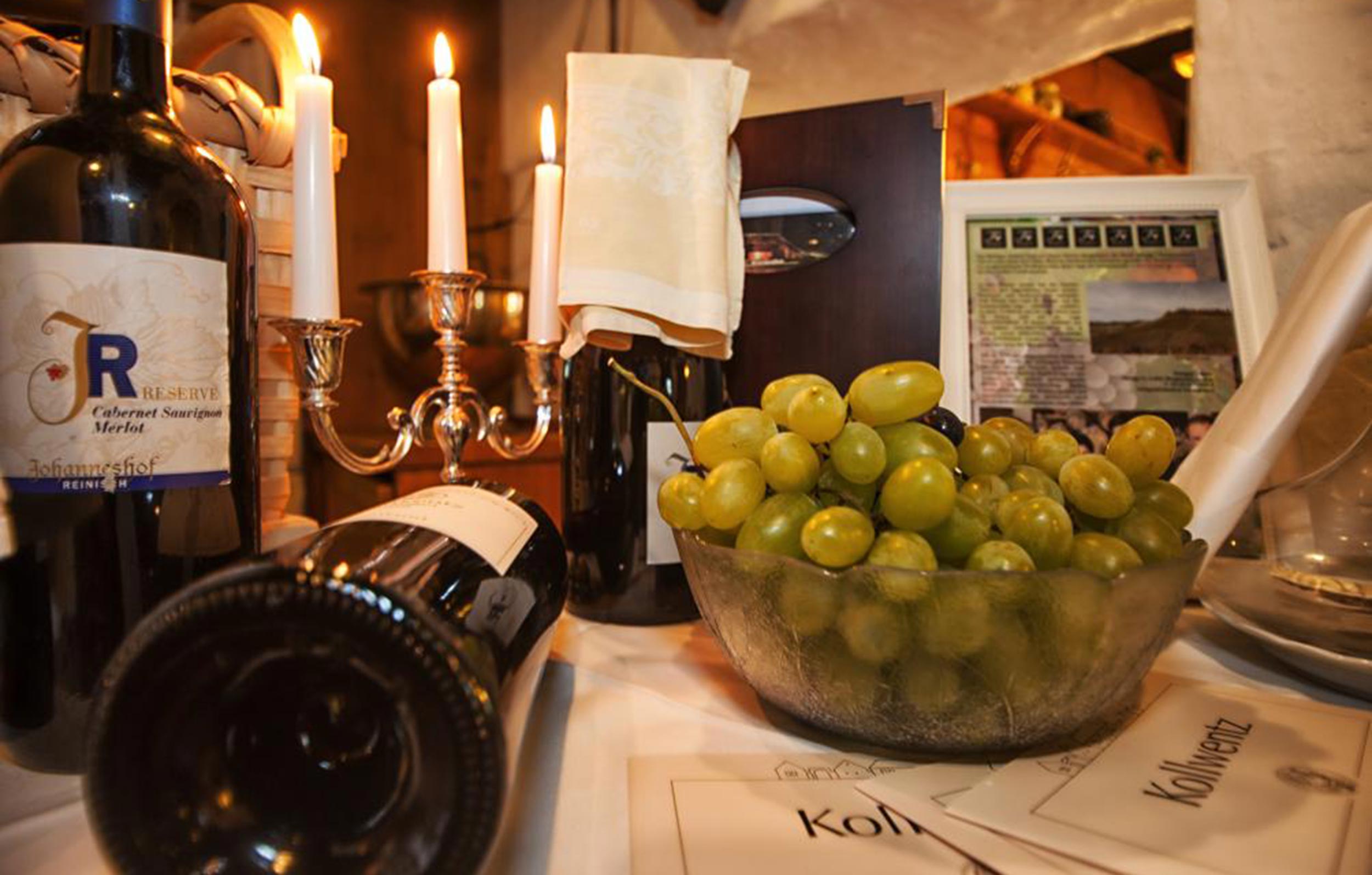 Grapes are in a bowl, next to them are some wine bottles, the picture is completed by a candlestick and a dish.